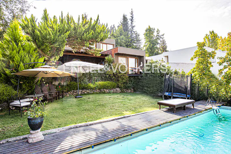 Backyard view of a modern house with a pool, wooden deck, green lawn, trees, and patio furniture.