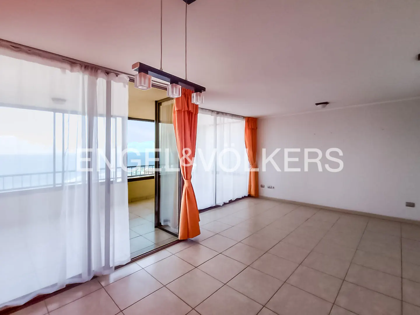 Empty living room with tiled floors, large windows, sheer white curtains, and bright orange drapes leading to a balcony.