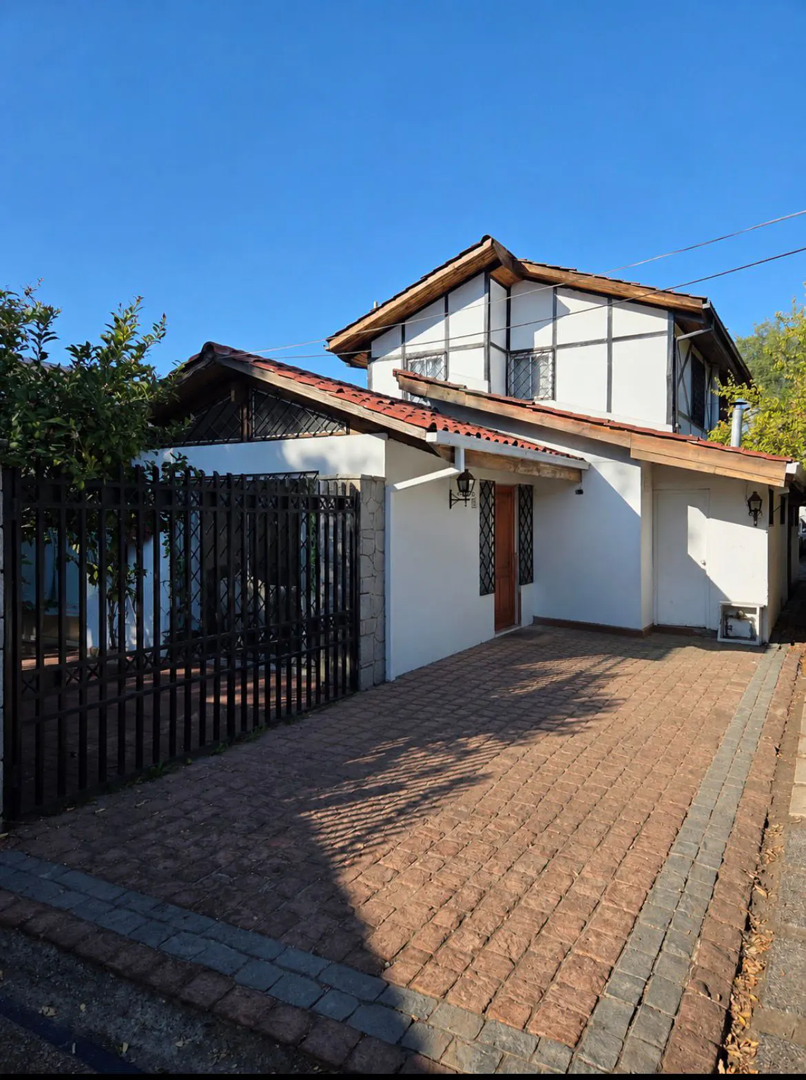 Two-story white house with dark timber framing, a red tile roof, and a paved driveway behind a black metal fence.