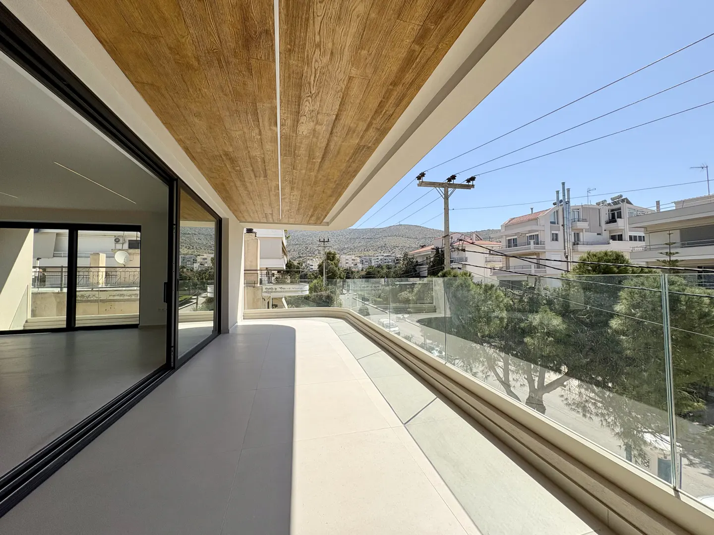 Balcony view with glass railing, light gray floor, and wood-look ceiling. Sliding glass doors lead to an empty room. Cityscape background.