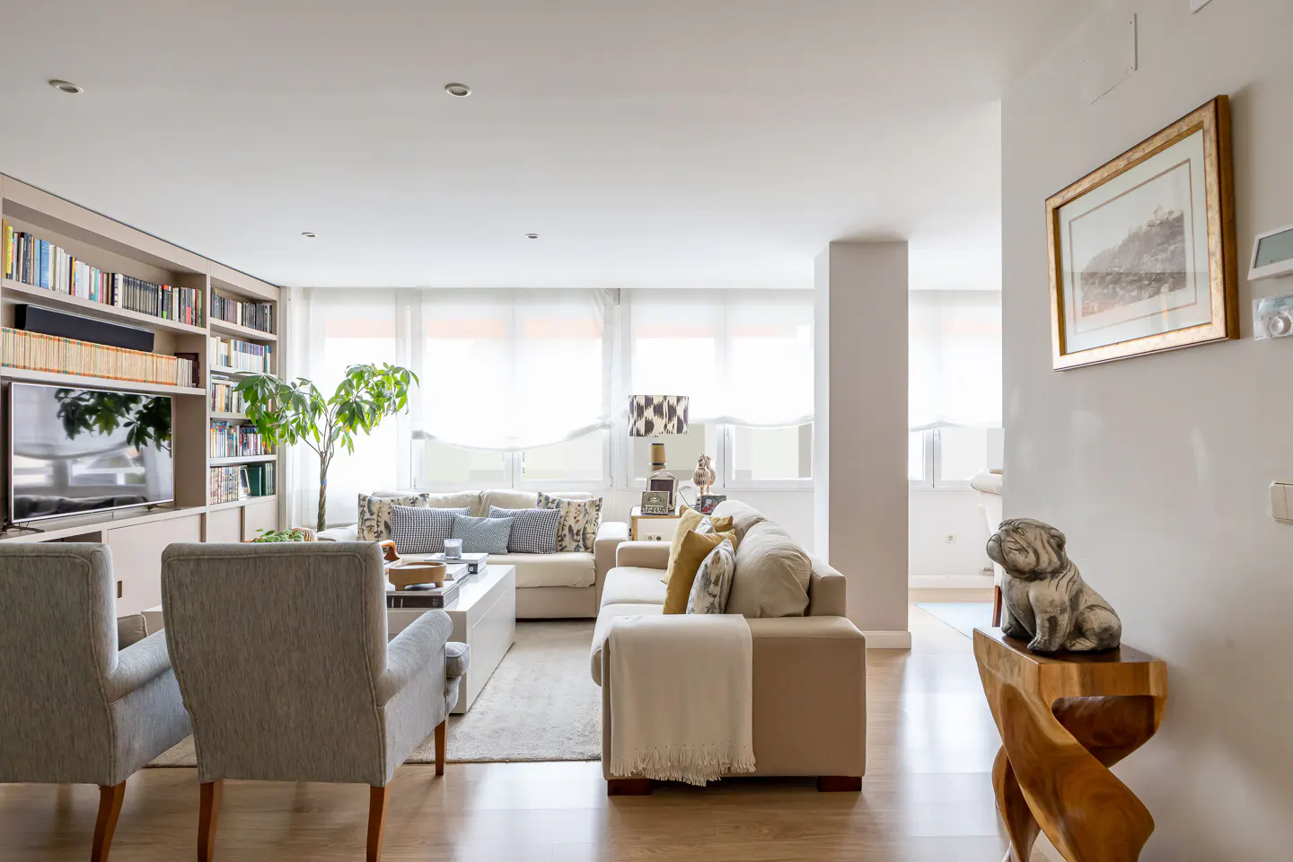 Bright living room with two sofas, two chairs, a bookshelf, and a wooden dog sculpture on a wooden table.