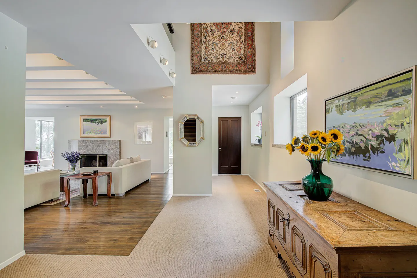Bright, open-concept home foyer with a wooden chest, sunflowers, and a painting. Living room with a fireplace is visible.