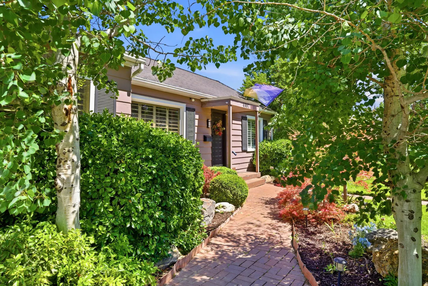 A brown house with black shutters and a brick walkway is surrounded by green trees and bushes.