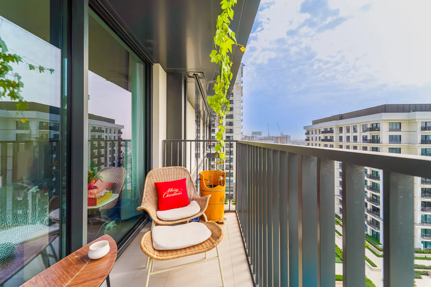 Balcony view with wicker chair, red pillow, and city skyline. Green vines hang from the ceiling. Metal railing and glass doors.