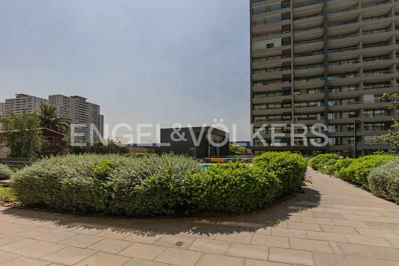 Exterior view of a tall apartment building with a pool, bushes, and a stone walkway. The Engel & Völkers logo is visible in the background.