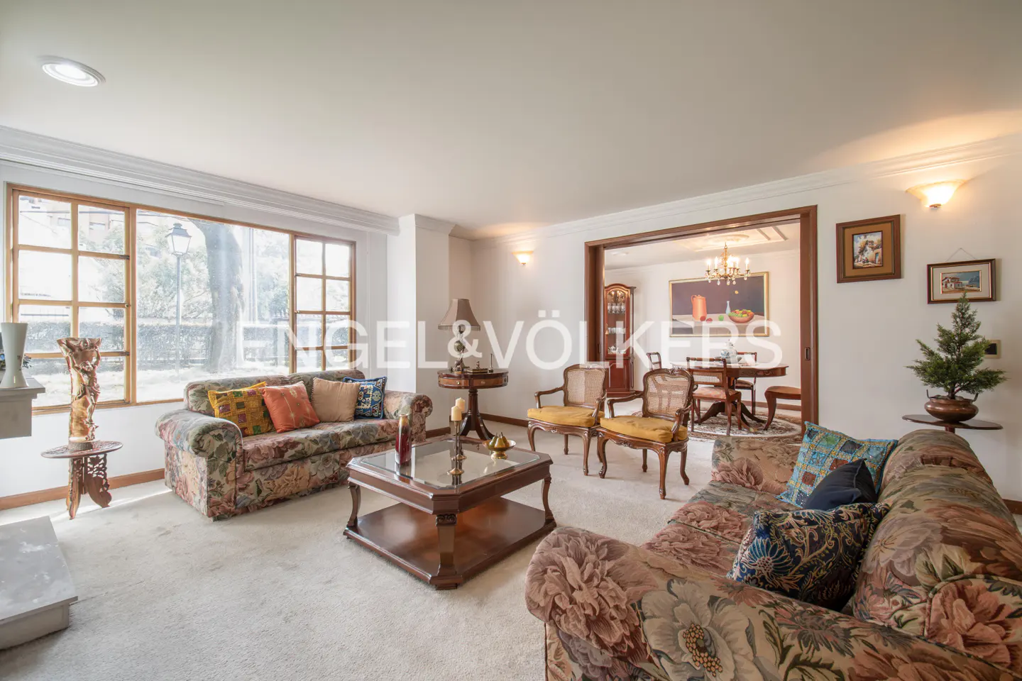 Living room with floral sofas, wood table, and chairs. Dining room visible through an archway. Large window with natural light.