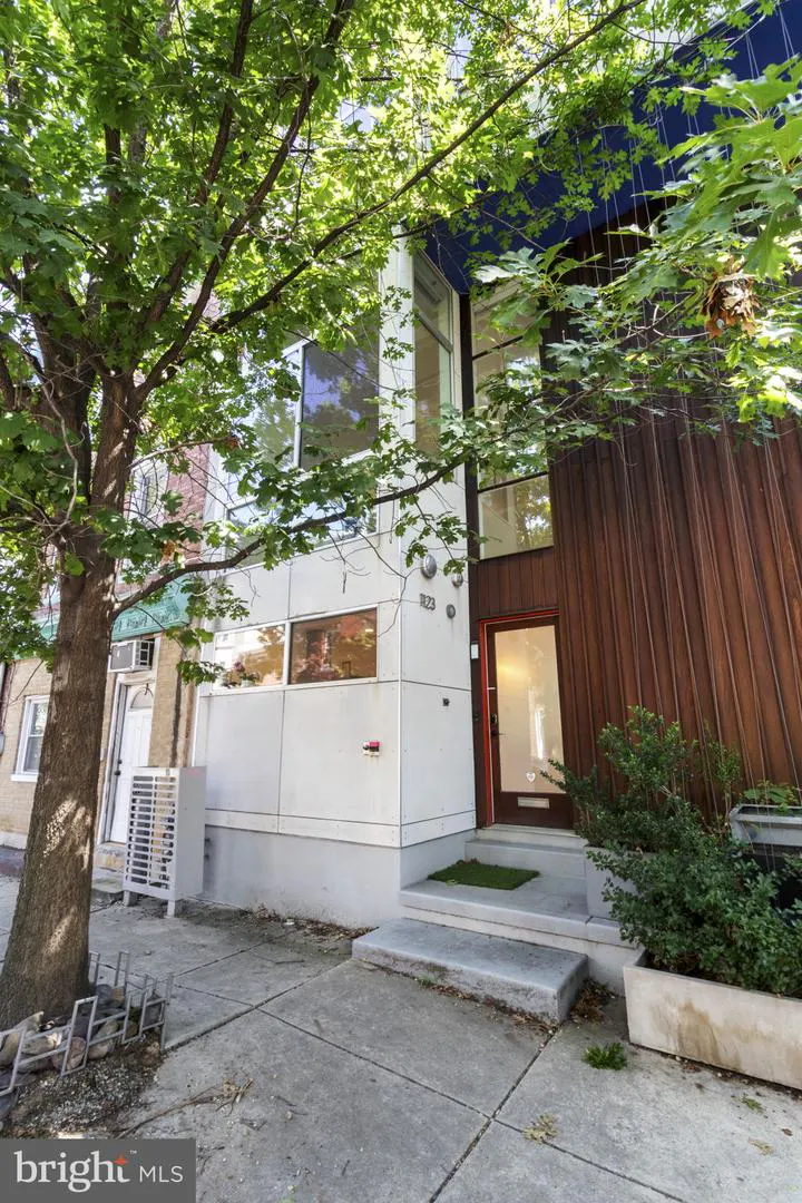 Modern two-story house with white and brown wood paneling, large windows, and a red-framed door. A tree shades the front.