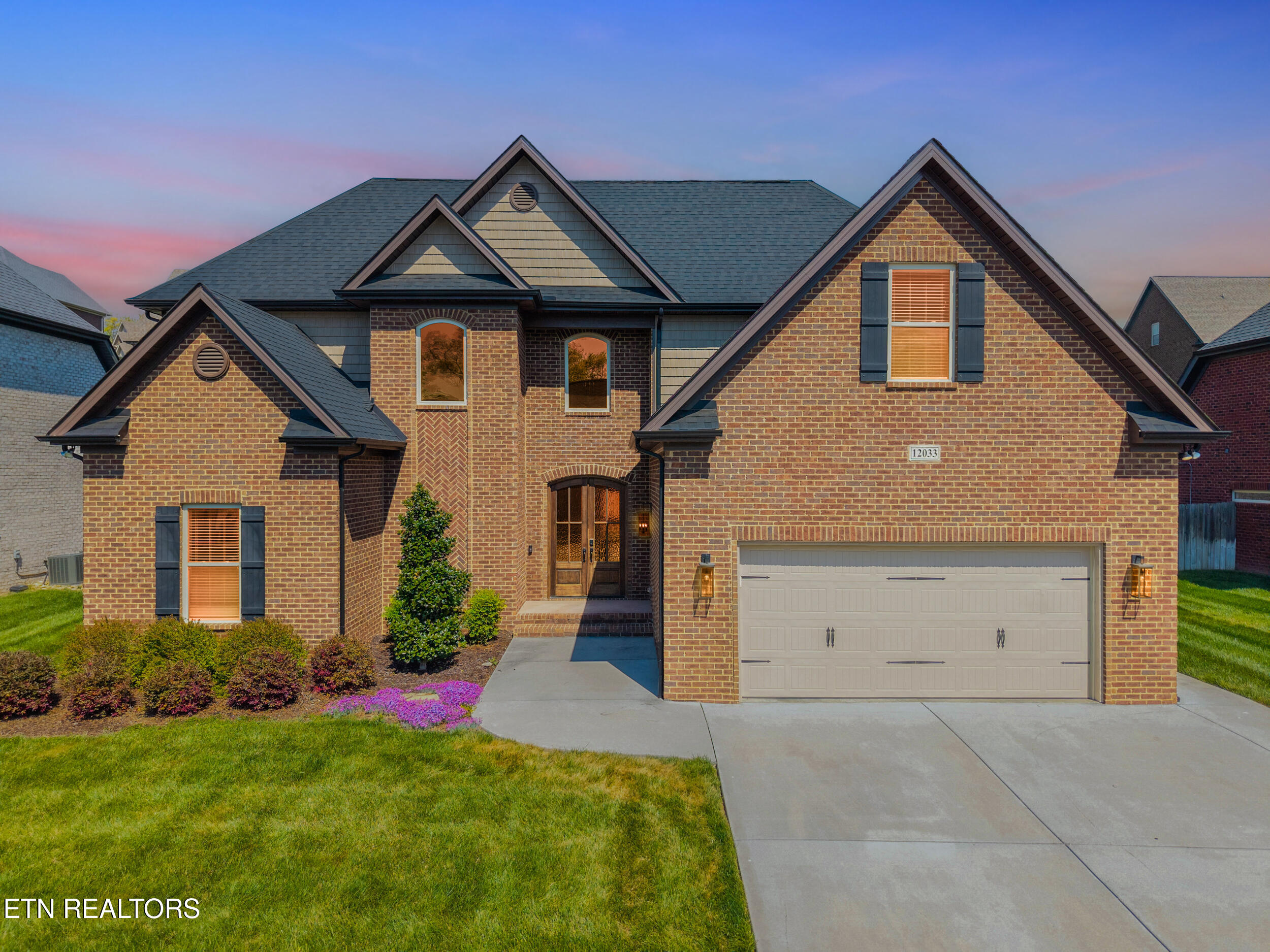 Two-story brick house with a dark roof, black shutters, and a two-car garage. Green lawn and purple flowers in front.
