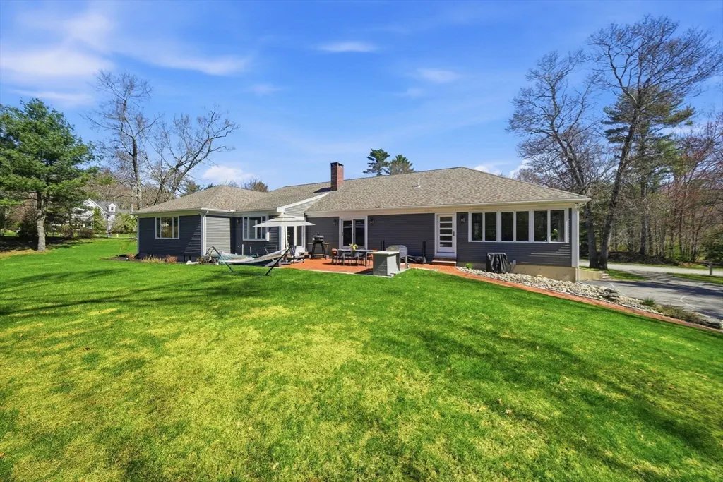 Backyard view of a gray house with a brick patio, lawn, and trees under a blue sky. Patio furniture and a hammock are visible.