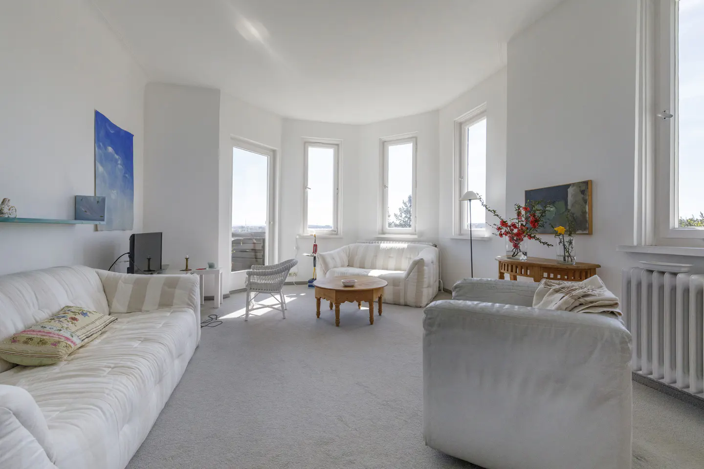 Bright, airy living room with white sofas, chair, and walls. Large windows offer a view. A round wooden table sits in the center.