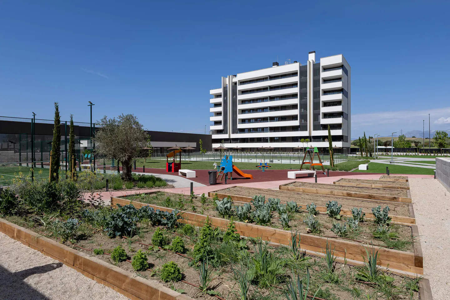 Apartment building with a community garden and playground. The building is white with black accents. The sky is blue.