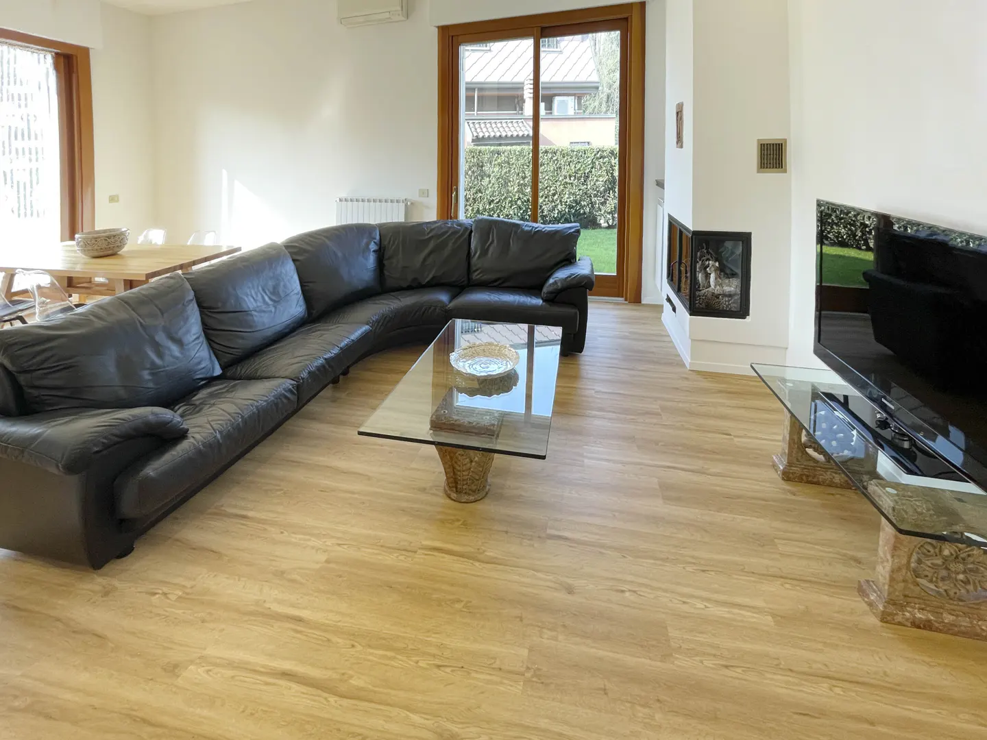 Bright living room with a black leather sectional sofa, glass coffee table, and a TV on a glass stand. A fireplace and a sliding glass door are visible.