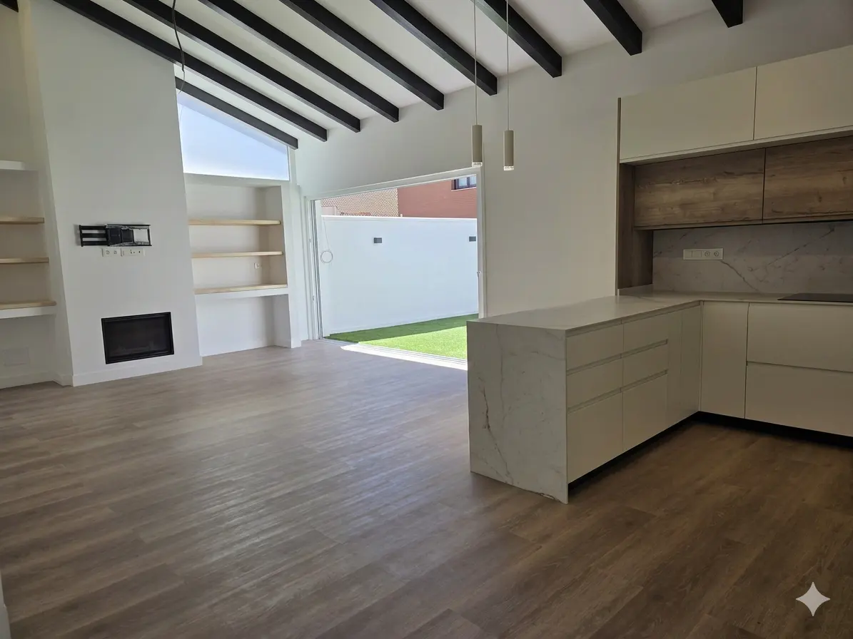 Bright, modern open-plan living space with wood floors, white walls, and black ceiling beams. Kitchen island and cabinets visible. Patio doors open to a green lawn.