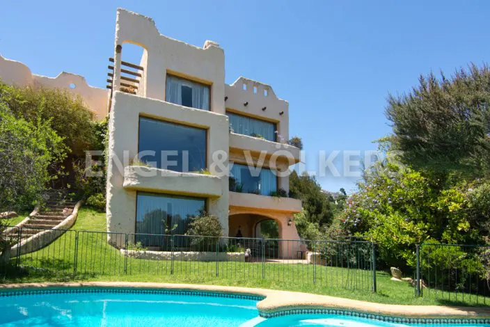 Exterior view of a multi-story beige house with large windows, a pool, and green lawn under a blue sky.