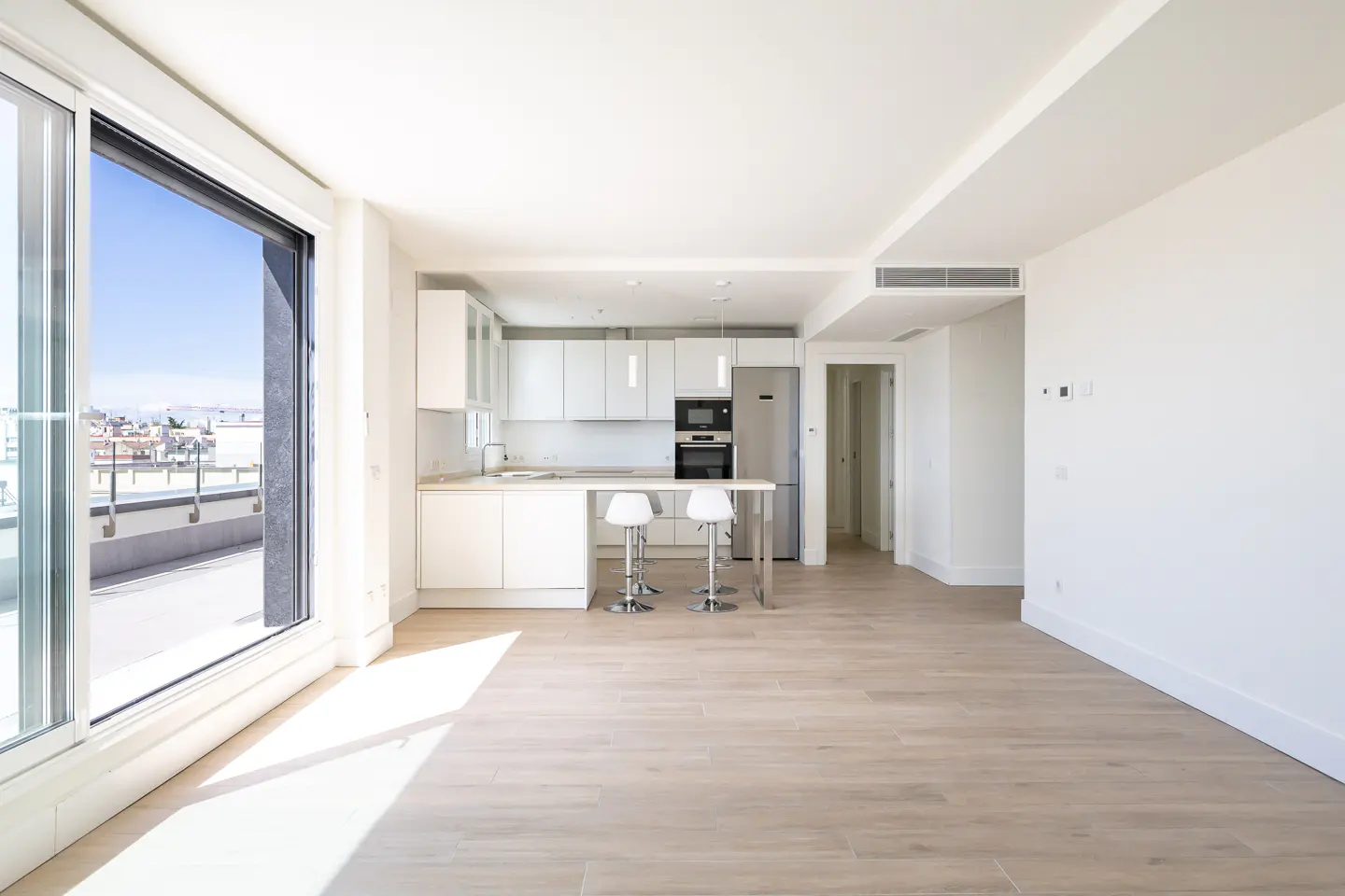 Bright, modern apartment with white walls, light wood floors, and a white kitchen with stainless steel appliances. Sliding glass doors open to a balcony.
