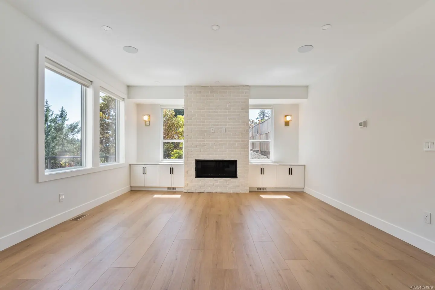 Bright, empty living room with light wood floors, white walls, and a white brick fireplace. Windows flank the fireplace.