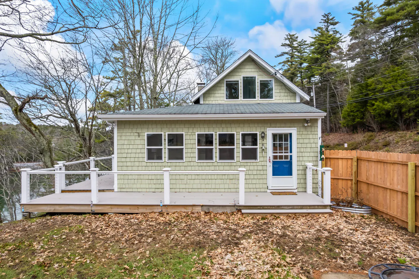 Exterior view of a light green shingle house with a blue door and a deck overlooking the water.