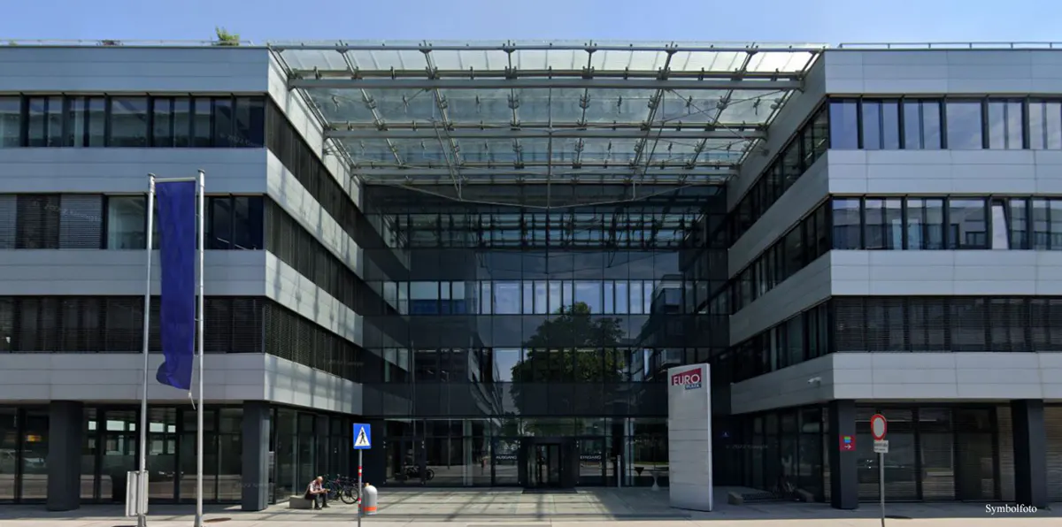 Modern office building with glass entrance and blue flag. Pedestrians and street signs are visible.