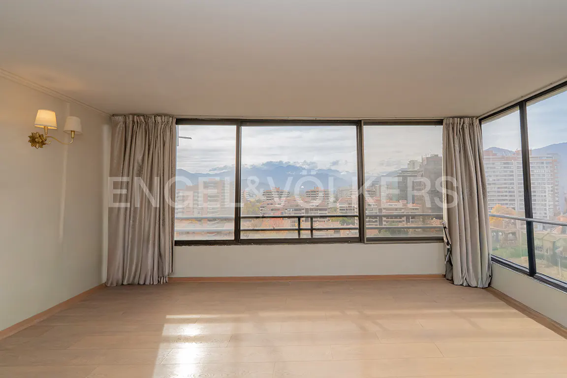 Empty room with light wood floors, large windows showing city and mountain views, and beige curtains.