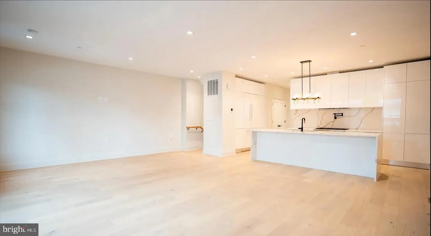 Bright, open-concept kitchen and living room with light wood floors and white walls. The kitchen features white cabinets and a marble backsplash.