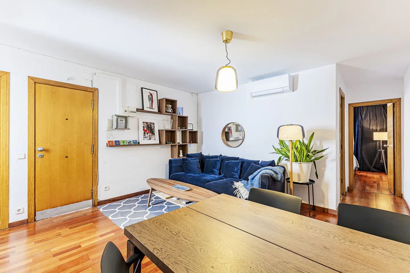 Bright living room with wood floors, a blue sofa, a wood table, and white walls with shelves and decor.