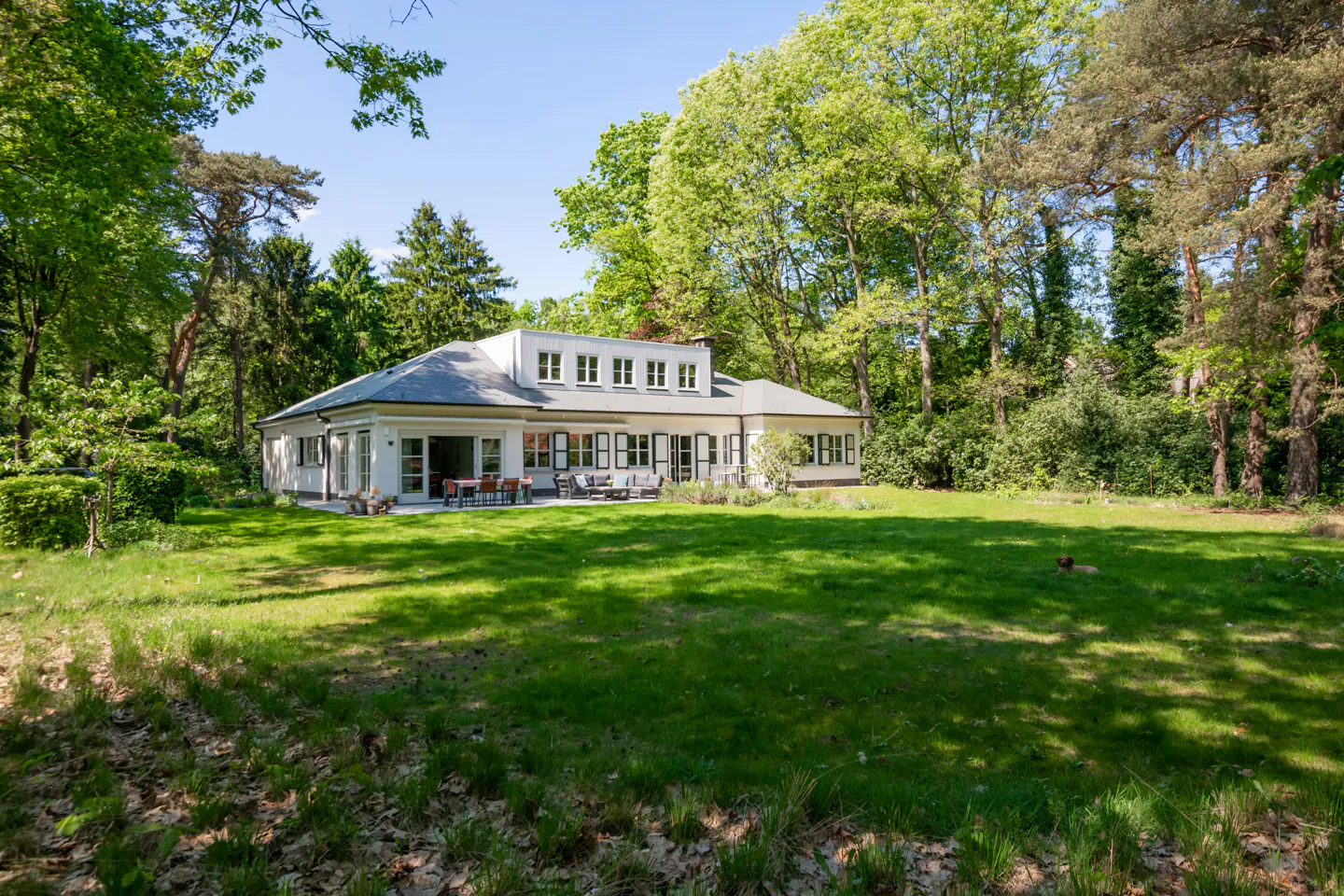 A white house with a gray roof sits on a green lawn surrounded by trees. Patio furniture is visible on the back patio.