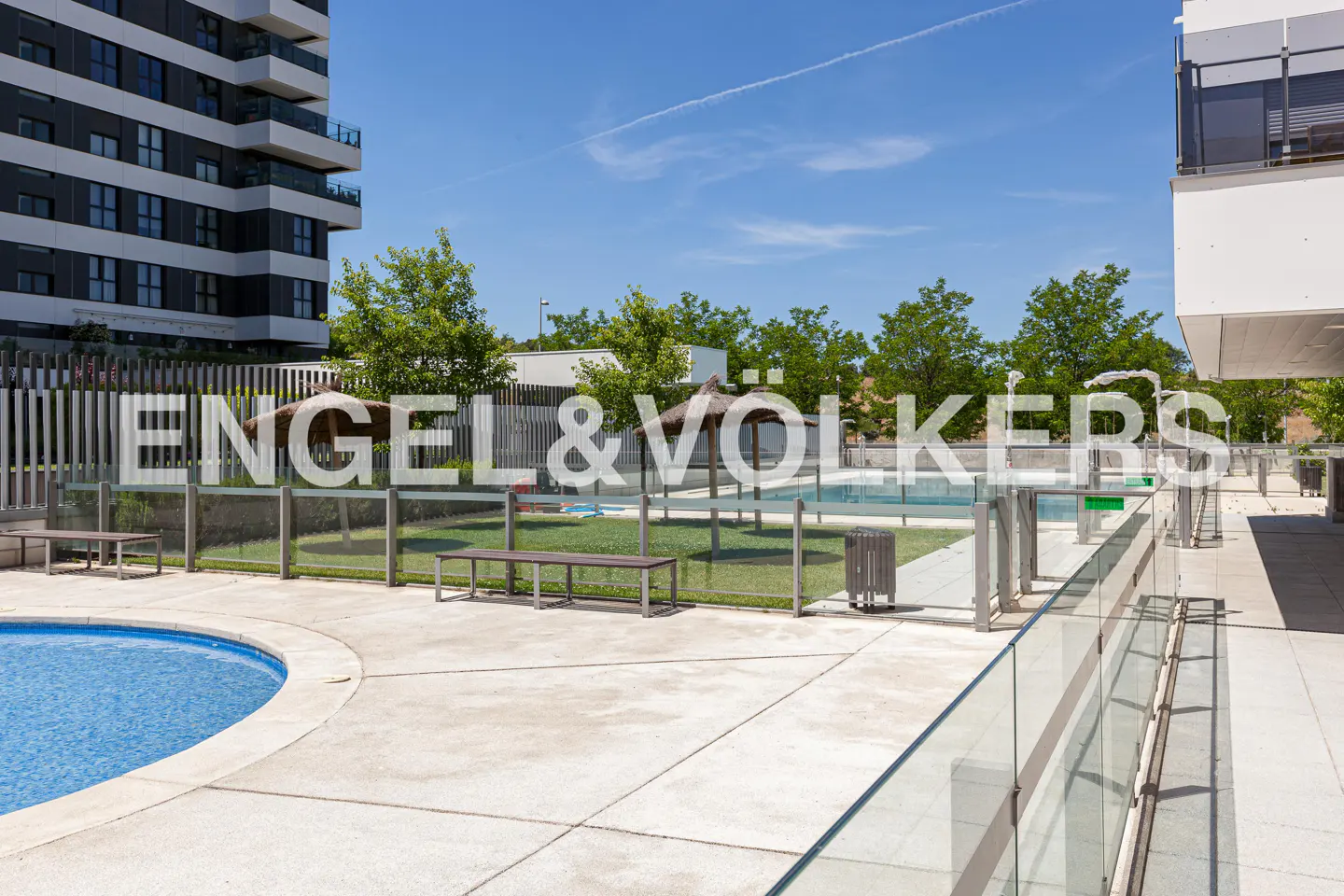Outdoor pool area with lounge chairs, umbrellas, and a modern apartment building in the background on a sunny day.