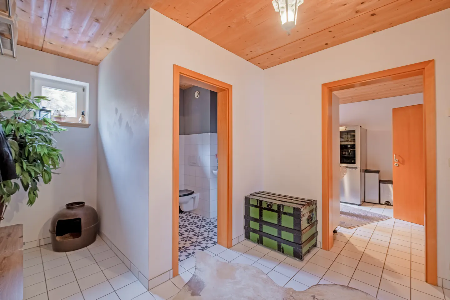 Hallway with white tile floor, wood ceiling, and orange door frames. A green chest sits near a bathroom entrance. A plant is near a window.