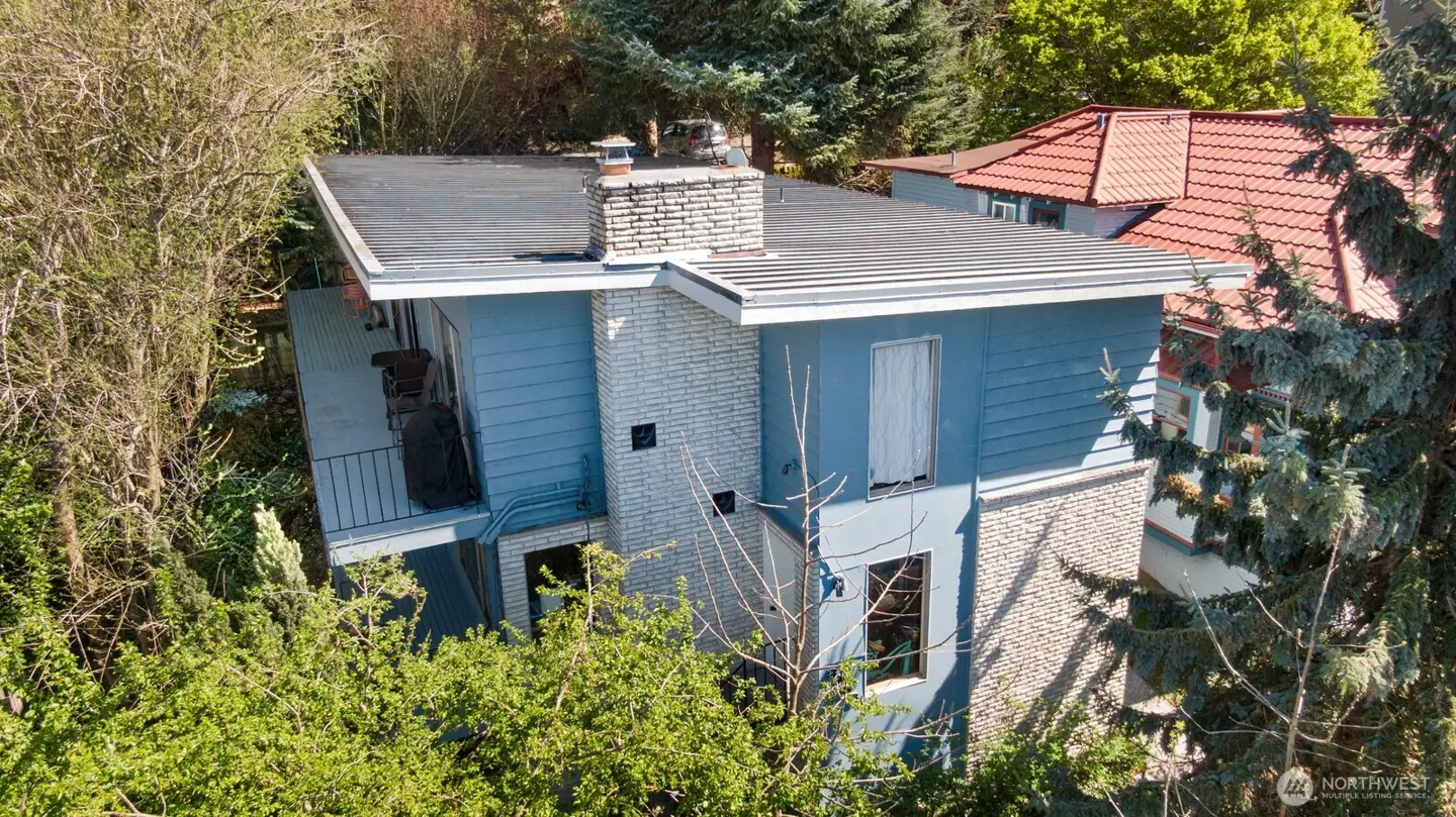 Aerial view of a two-story blue house with a flat black roof, brick chimney, and a small balcony surrounded by lush green trees.