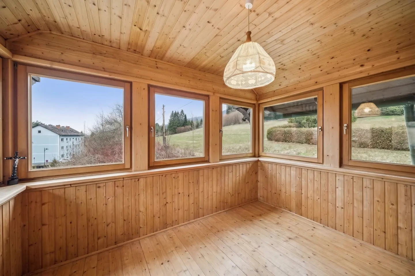 A sunroom with wood paneled walls, ceiling, and floor. Large windows offer views of the outside. A white pendant light hangs from the ceiling.