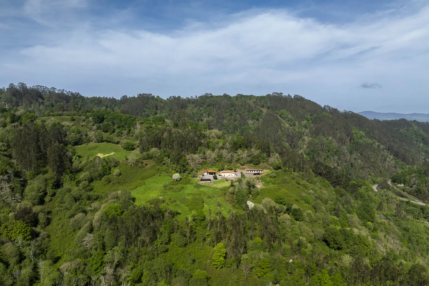 Aerial view of a house with a red roof on a green, tree-covered hillside under a blue sky.