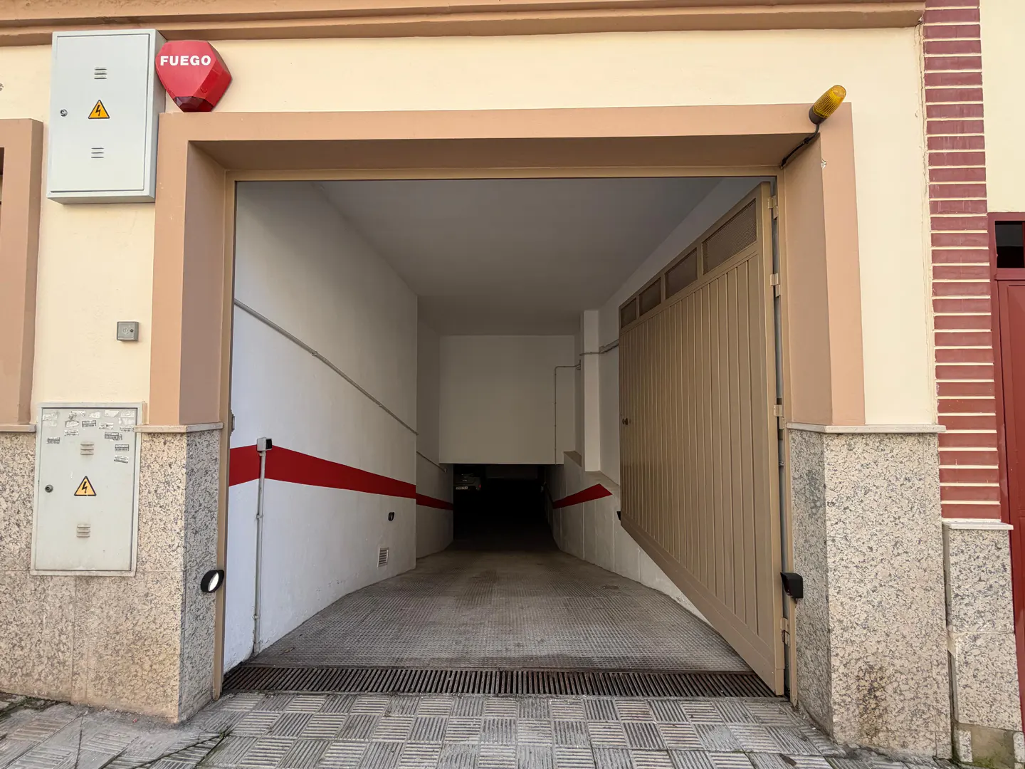 Open garage entrance with a beige metal gate, leading to a dark, downward-sloping ramp. Red stripe on the white wall. "Fuego" fire alarm box on the wall.