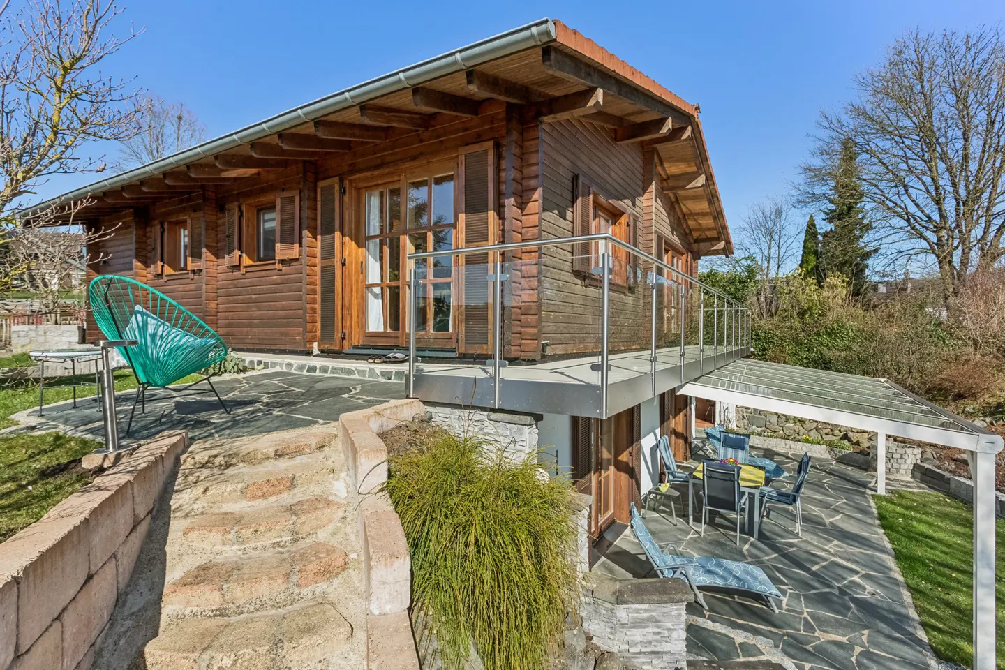 Exterior view of a two-story log cabin with a stone patio, outdoor furniture, and a green chair.