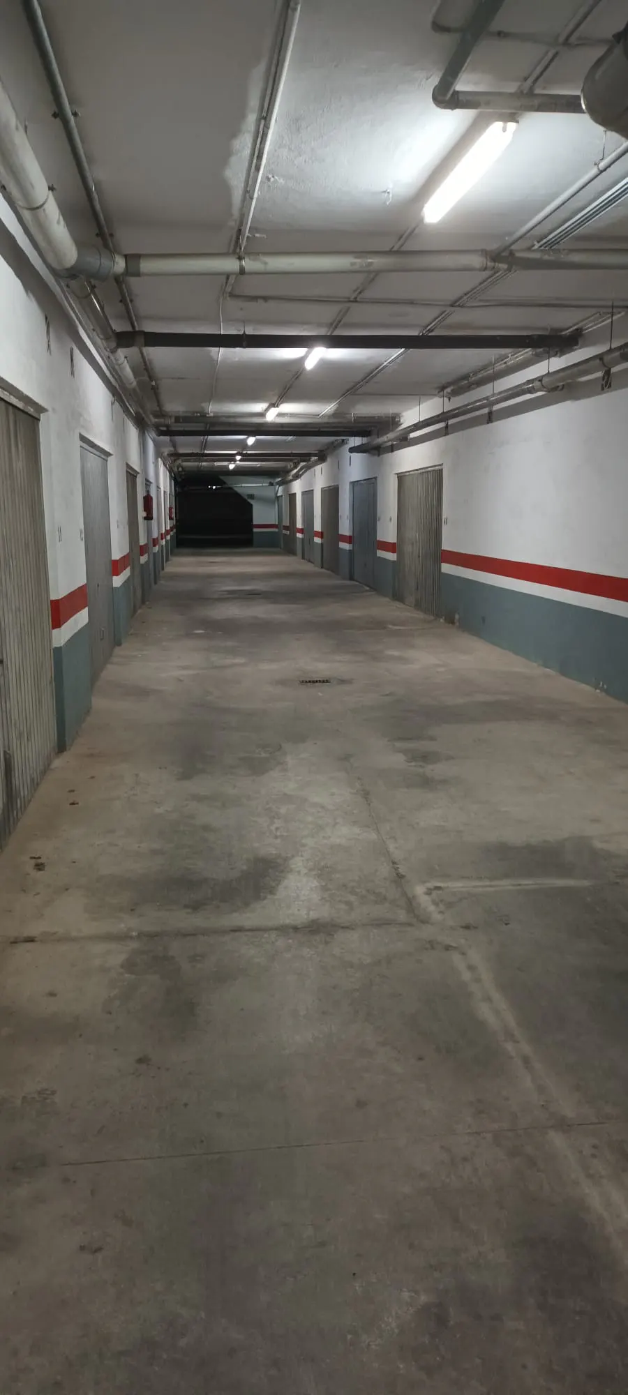 Empty underground parking garage with concrete floors, white walls, red stripes, and overhead fluorescent lighting.