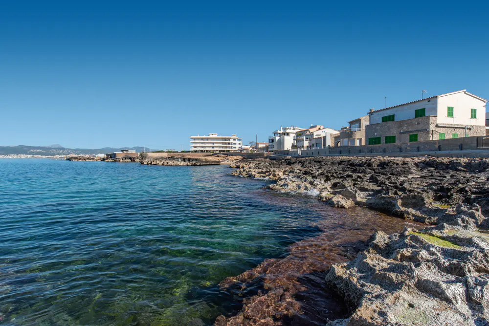 Rocky shoreline with turquoise water and white buildings under a clear blue sky.