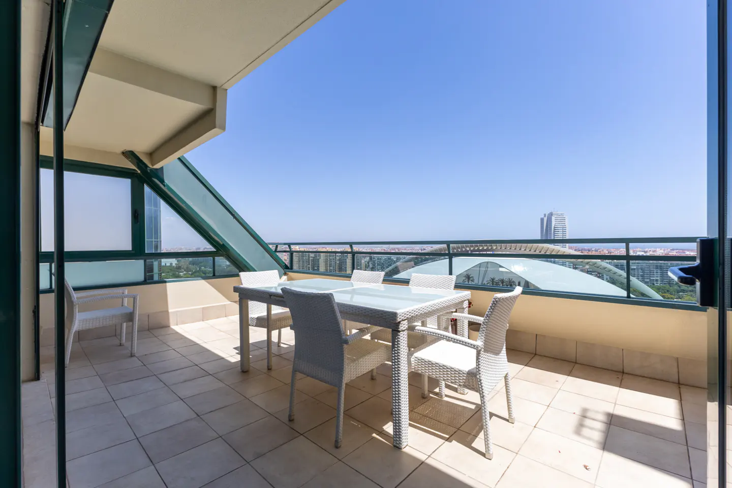 Spacious balcony with white wicker dining set and chairs, overlooking a city skyline under a clear blue sky.