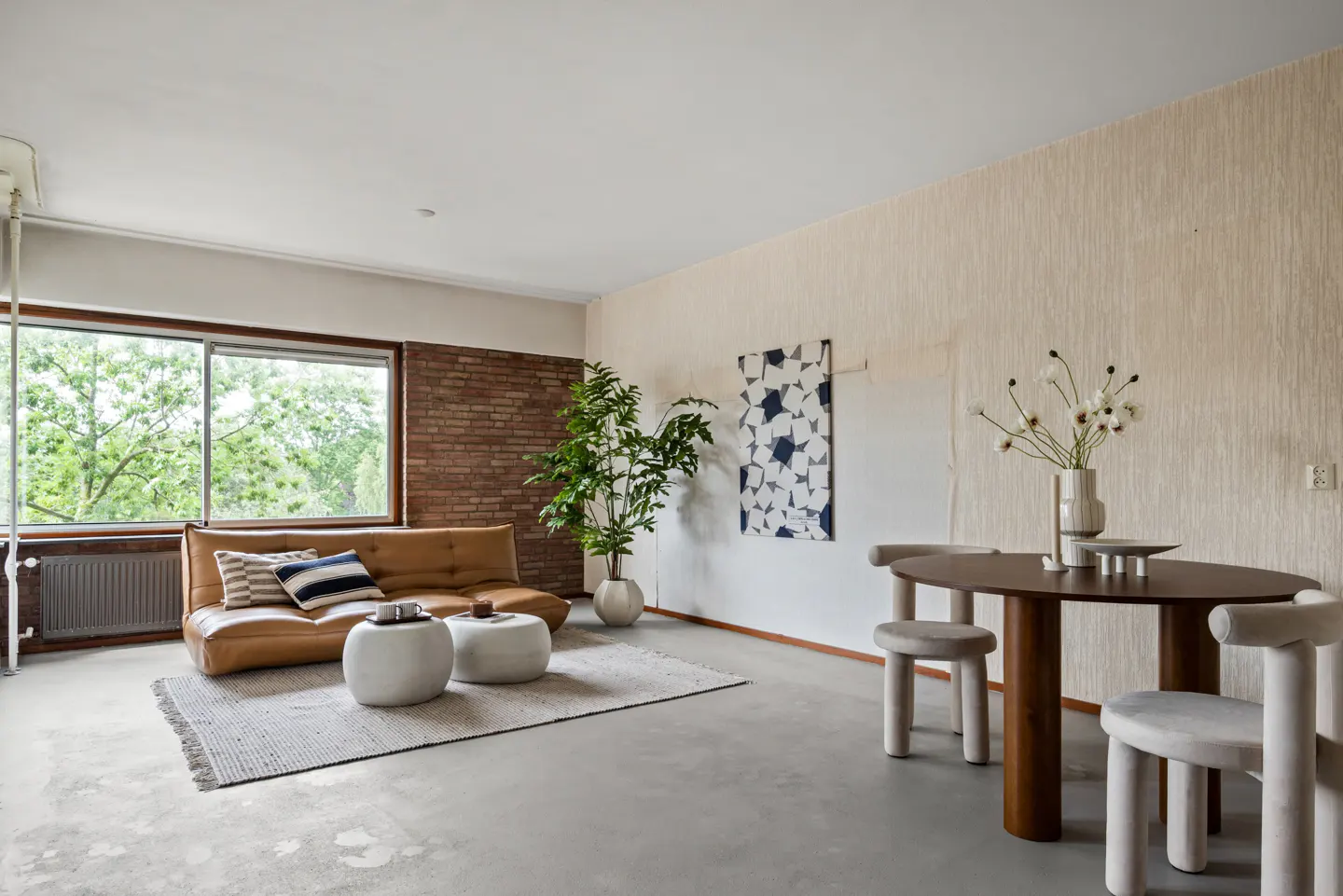 Modern living room with a tan leather sofa, two round white ottomans, a dining table, and a large indoor plant.