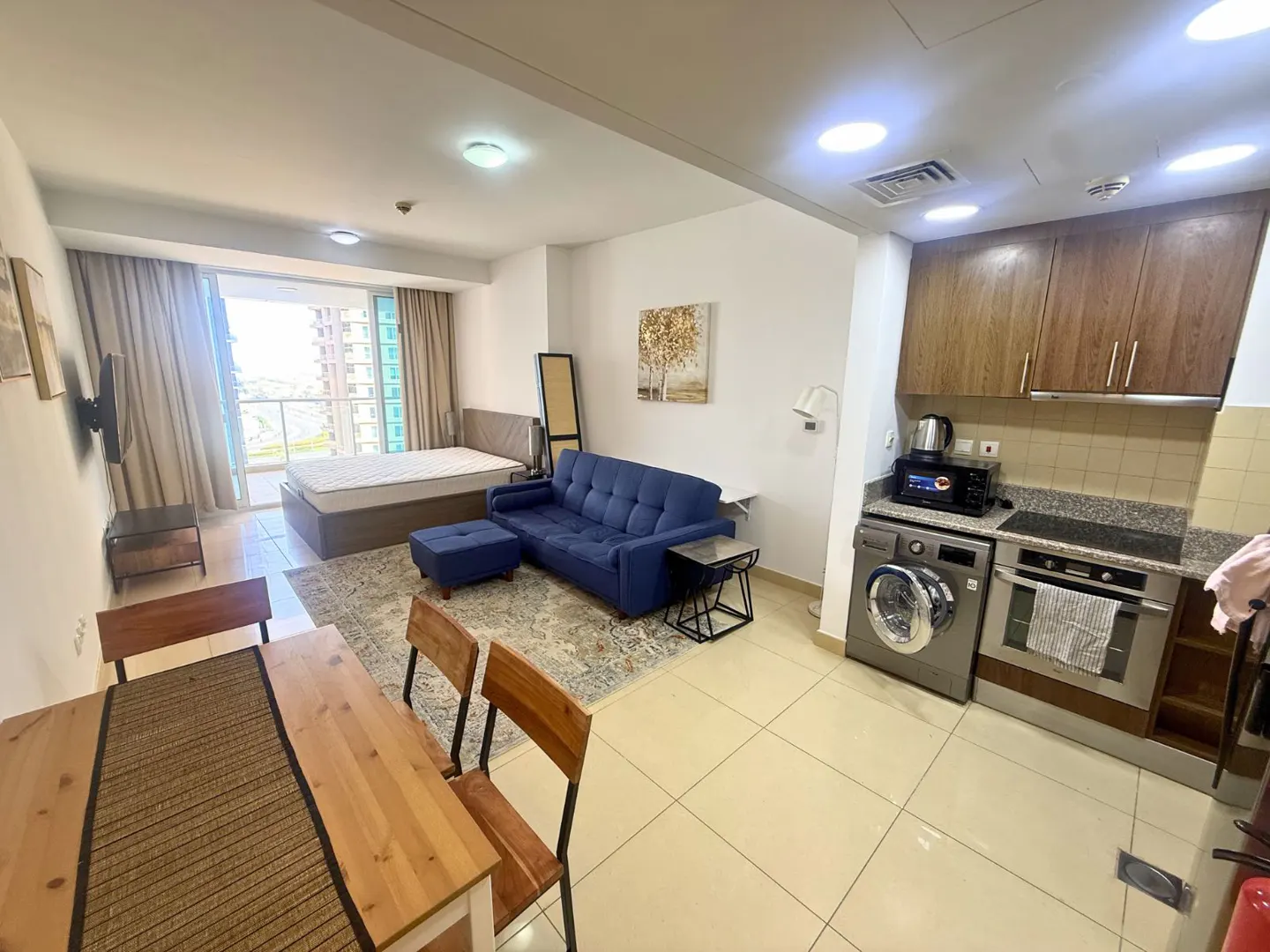 A wide shot of a furnished studio apartment with a bed, blue sofa, dining table, and kitchen area. Neutral tones with pops of blue.