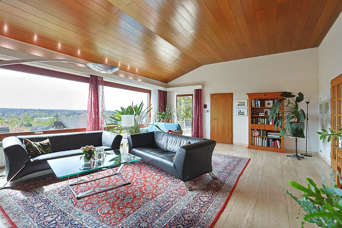 Living room with black leather sofas, glass coffee table, patterned rug, wooden ceiling, and large windows with a view.