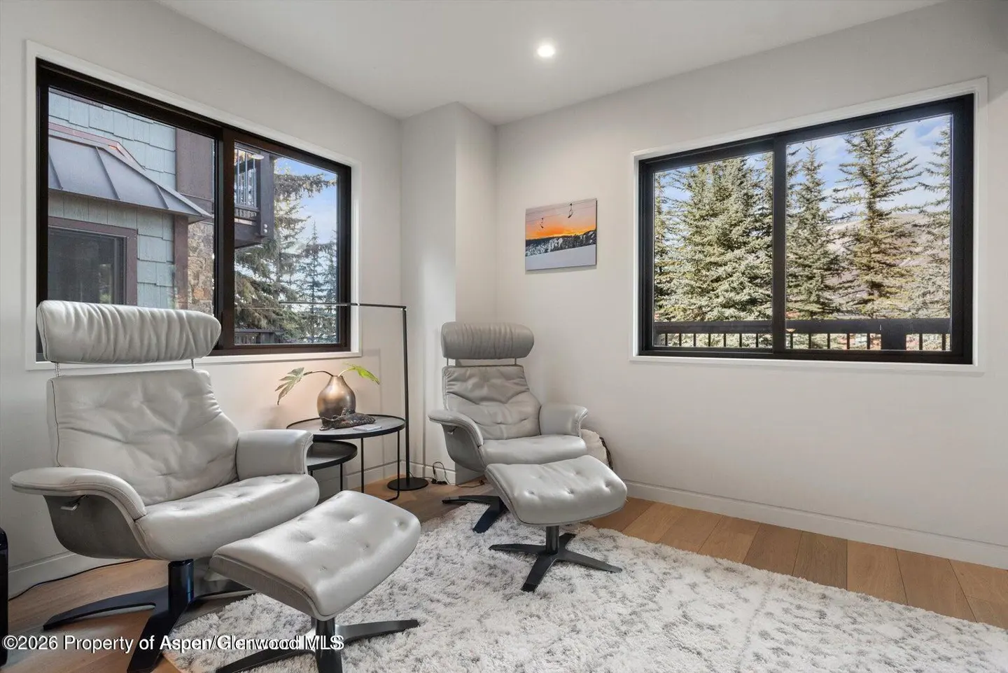 Bright room with two gray leather chairs, footrests, and black accents. Windows show trees and a building. A small table with a vase sits beside one chair.