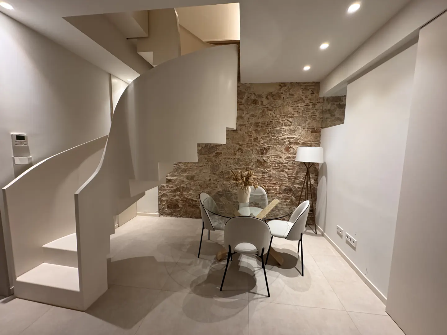 A modern interior with a white spiral staircase, stone wall, and a glass table with white chairs. A floor lamp stands near the wall.