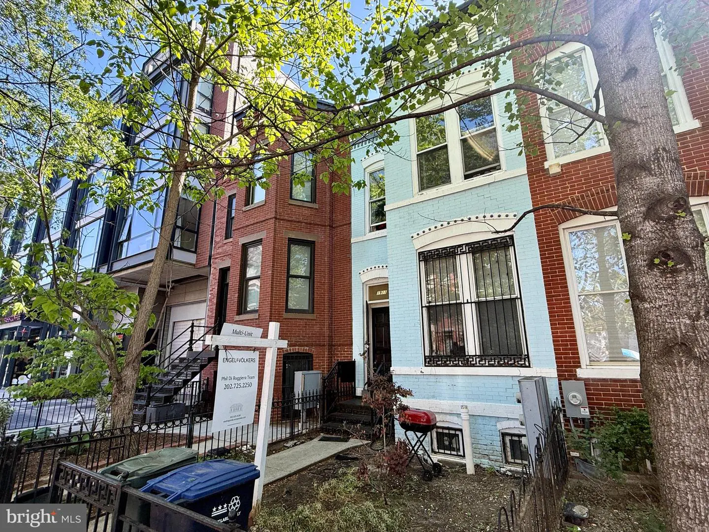 Exterior view of row houses, one light blue, one red brick, with a real estate sign in front.