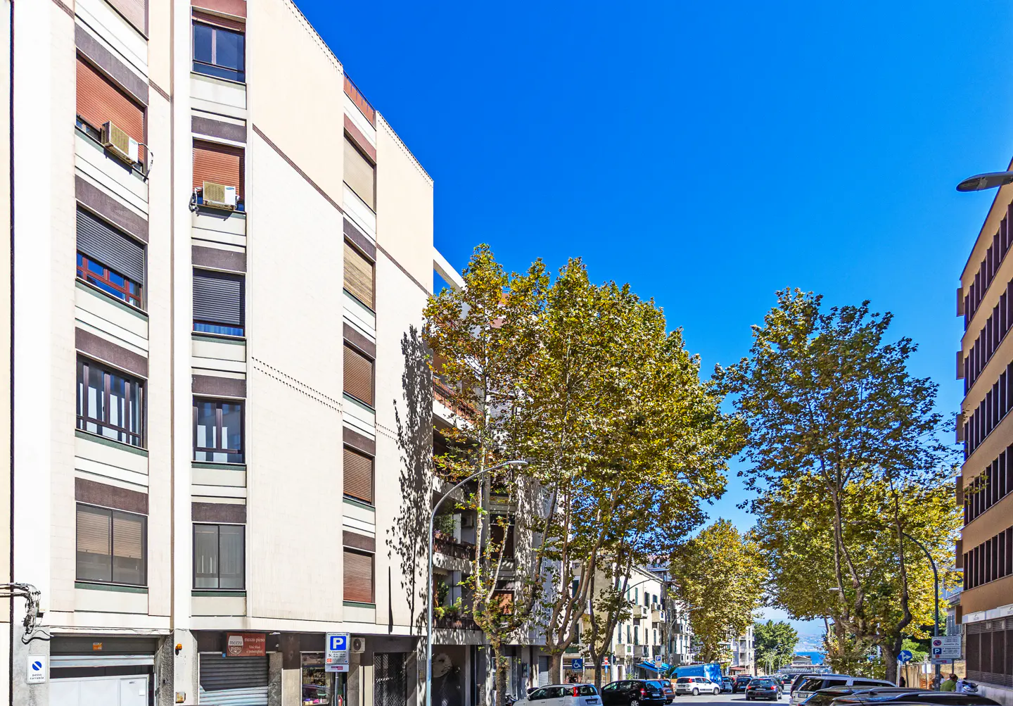 Street view of multi-story buildings with beige facades, brown window trim, and green trees under a clear blue sky. Cars are parked along the street.