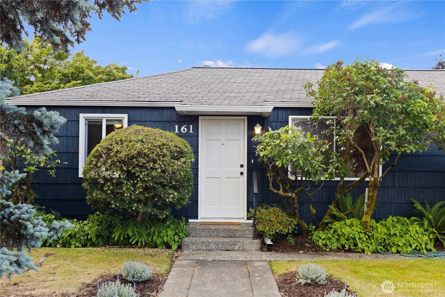 Single-story house with dark blue siding, white front door numbered 161, and lush green landscaping under a blue sky.