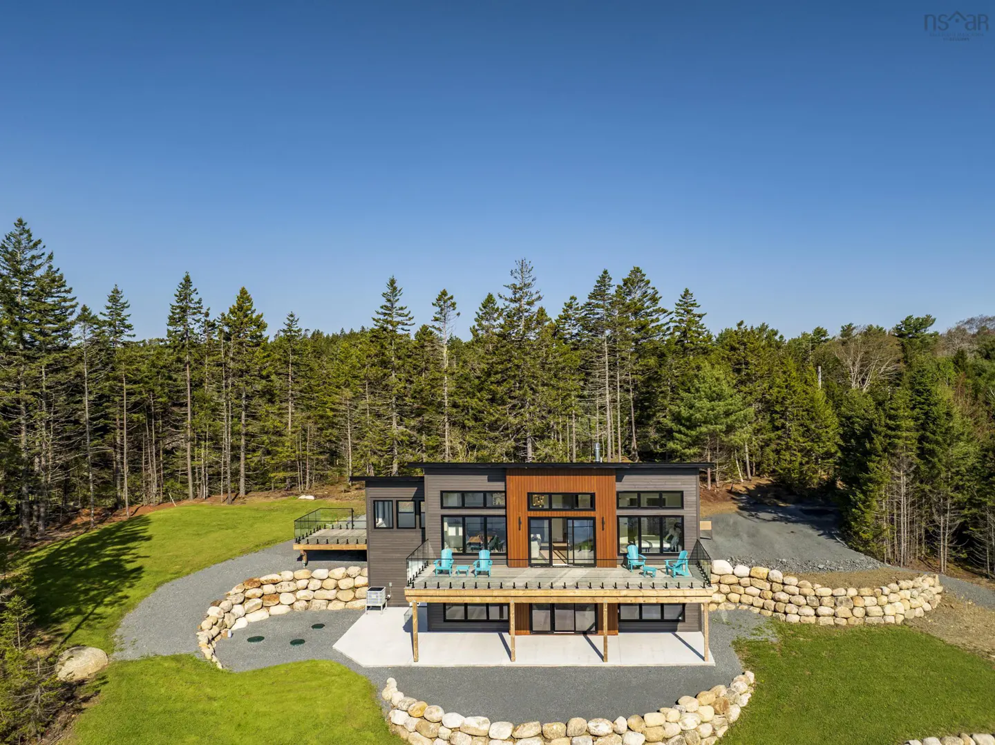Aerial view of a modern, gray house with a wood-accented facade, surrounded by a lush green lawn and a dense forest under a clear blue sky.