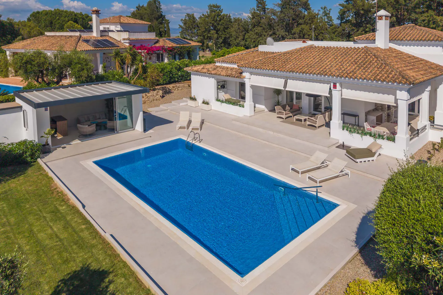 Aerial view of a white villa with a blue swimming pool, lounge chairs, and a covered patio. The villa has a terracotta roof and is surrounded by green trees.