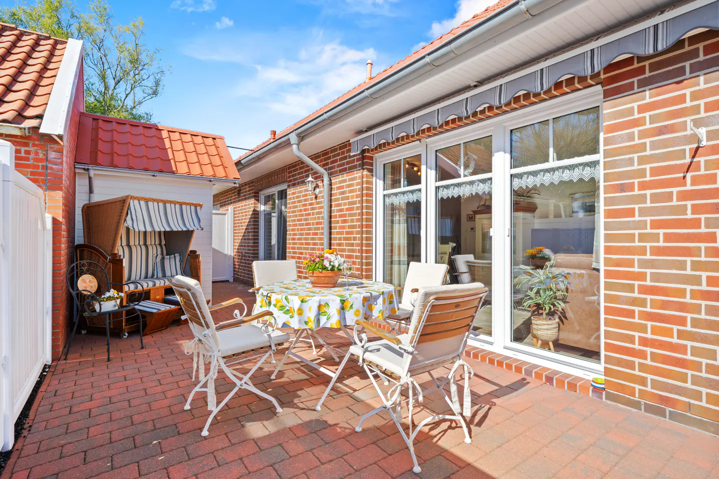 Brick patio with table, chairs, and a hooded beach chair. The house has red brick walls and a red tile roof. Blue sky.