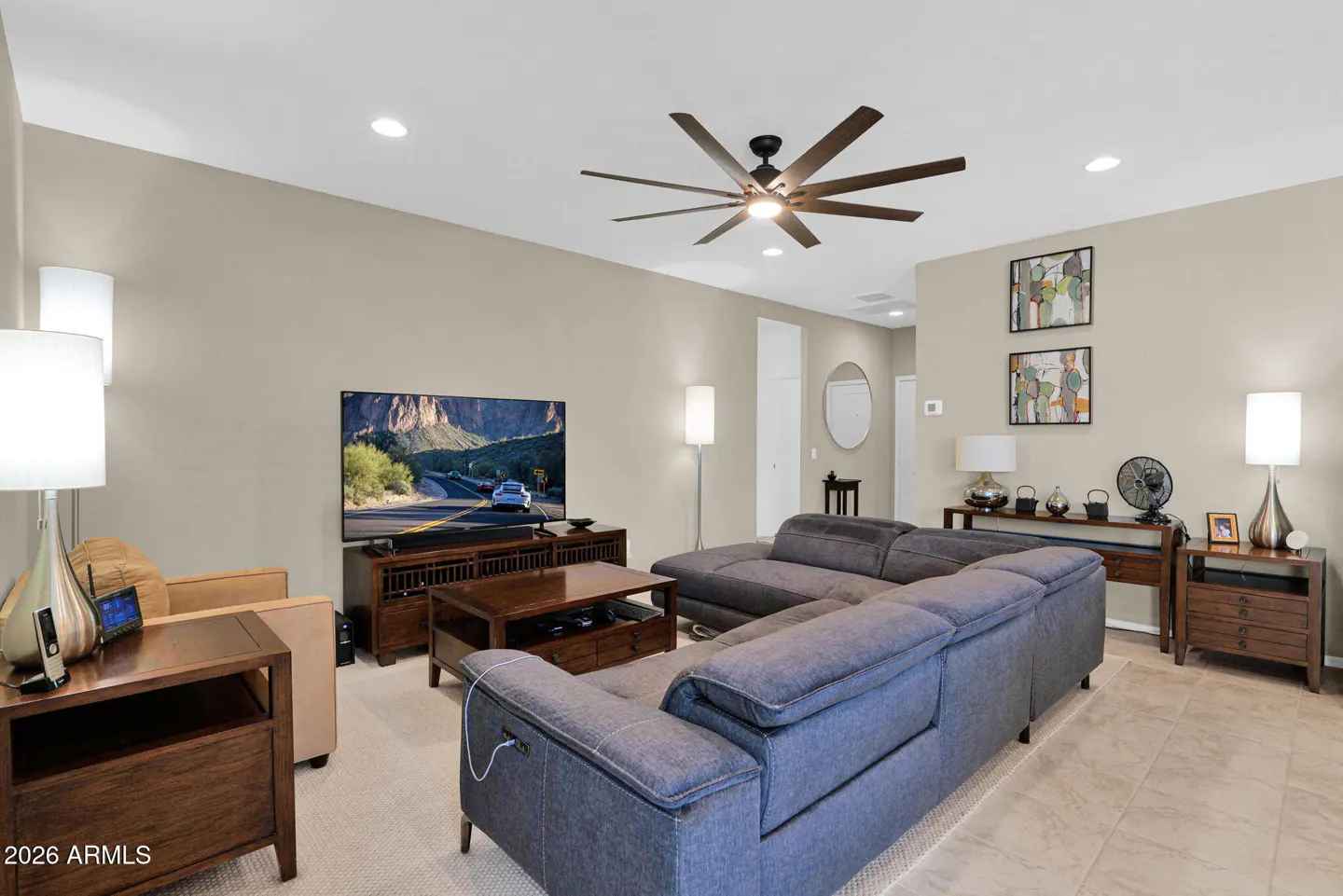 Living room with a gray sectional sofa, TV showing a mountain scene, and a brown ceiling fan.