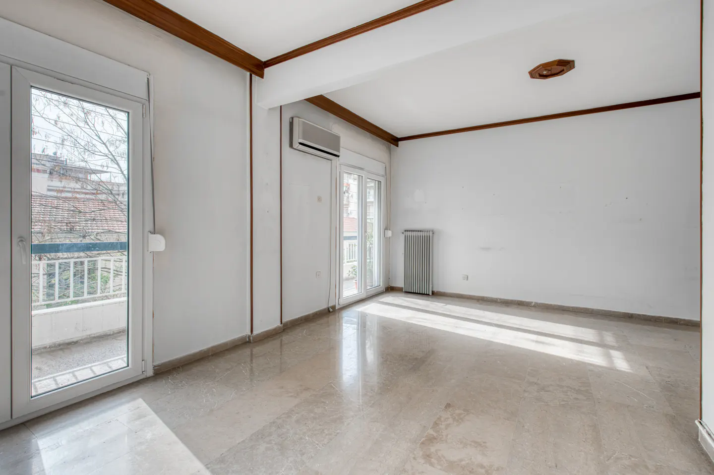 Bright, empty room with marble floors, white walls, and wood trim. Balcony doors let in sunlight. Radiator and AC unit visible.