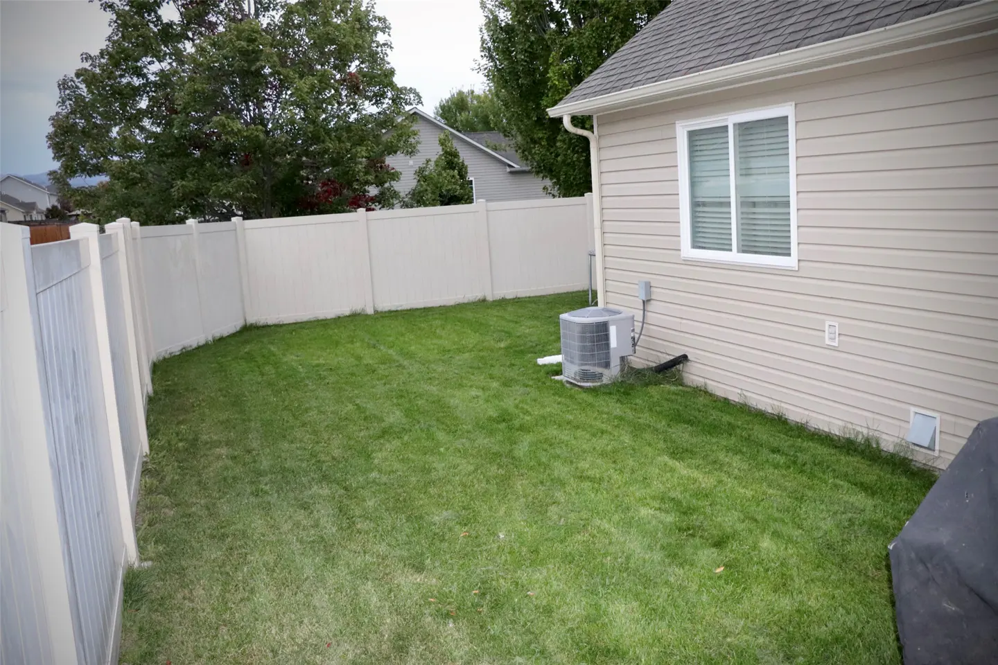 A fenced backyard with green grass, an AC unit, and a beige house with a white window.